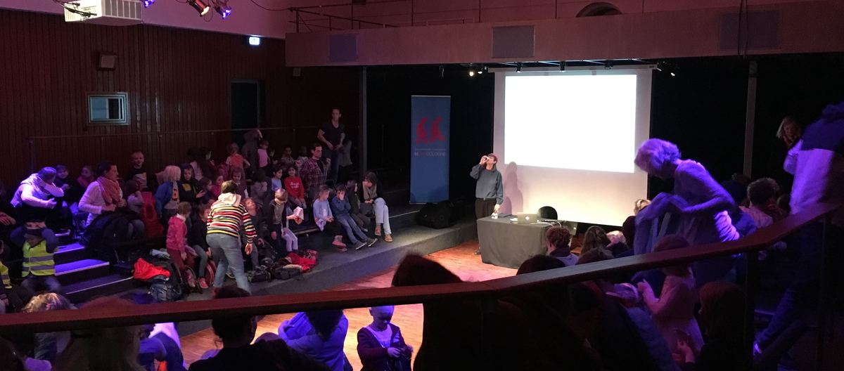 Ein voller Saal kurz vor der Lesung: Kinder sitzen auf den Stufen, vorn die Leinwand und der Lesetisch.