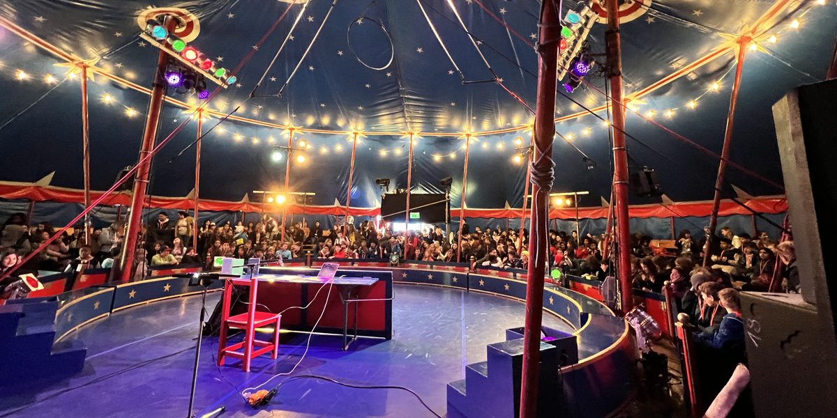 Volles Zirkuszelt in Hannover: Kuppel mit Sternen und Lichterkette, ringsum Publikum, in der Manege der Leseplatz mit rotem Stuhl, Tisch und Mikrofon.