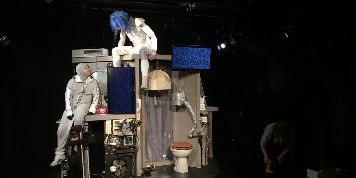 Szene aus „Zonka und Schlurch“: zwei Figuren in weißen Anzügen, eine mit blauer Perücke, auf einem Haus aus Waschmaschine, Klo und Fernsehern.