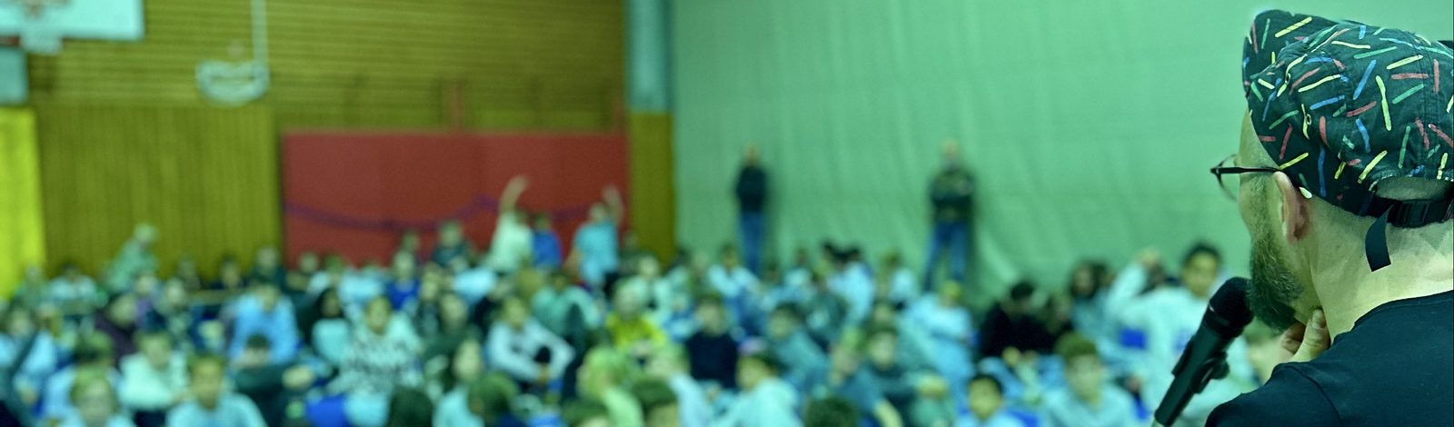 Finn-Ole Heinrich im Profil am Mikrofon, vor ihm eine volle Turnhalle mit Kindern auf Matten, einige melden sich.